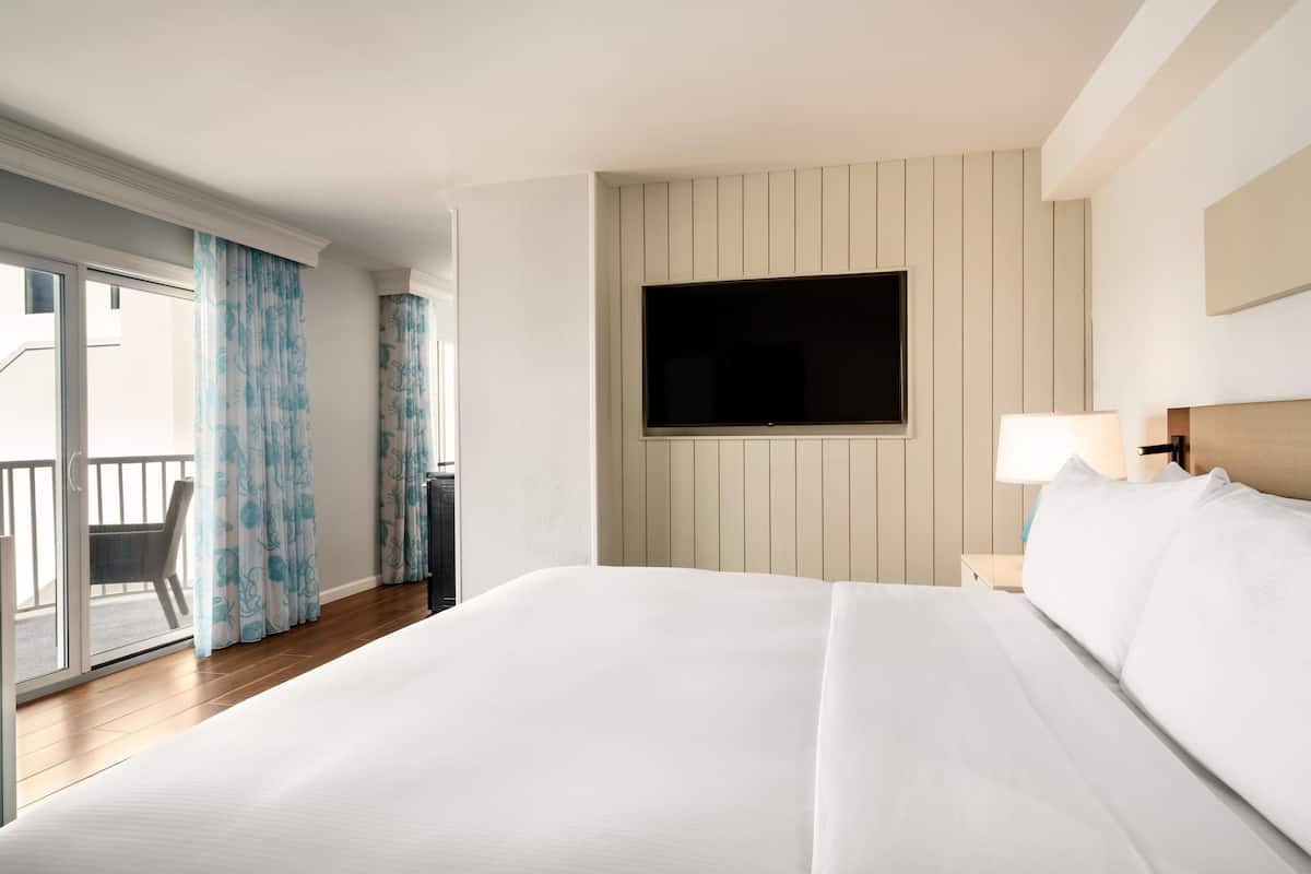 A hotel room with a large bed, a TV mounted on the wall, a nightstand with a lamp, and a balcony with curtains.
