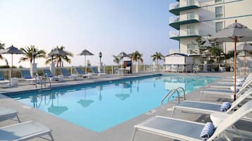 Zona de piscina con sillones de descanso, sombrillas y un edificio con balcones.