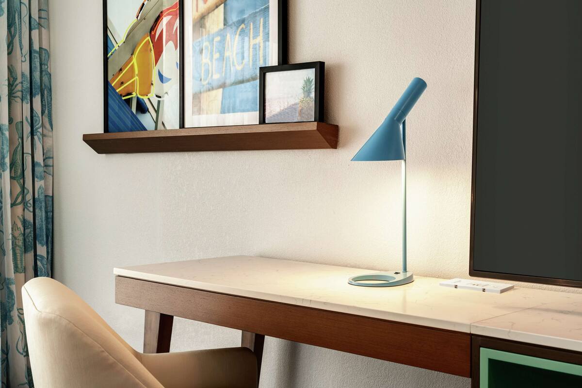 A hotel room with a desk, a chair, a blue desk lamp, a framed picture on the wall, and a window with curtains.
