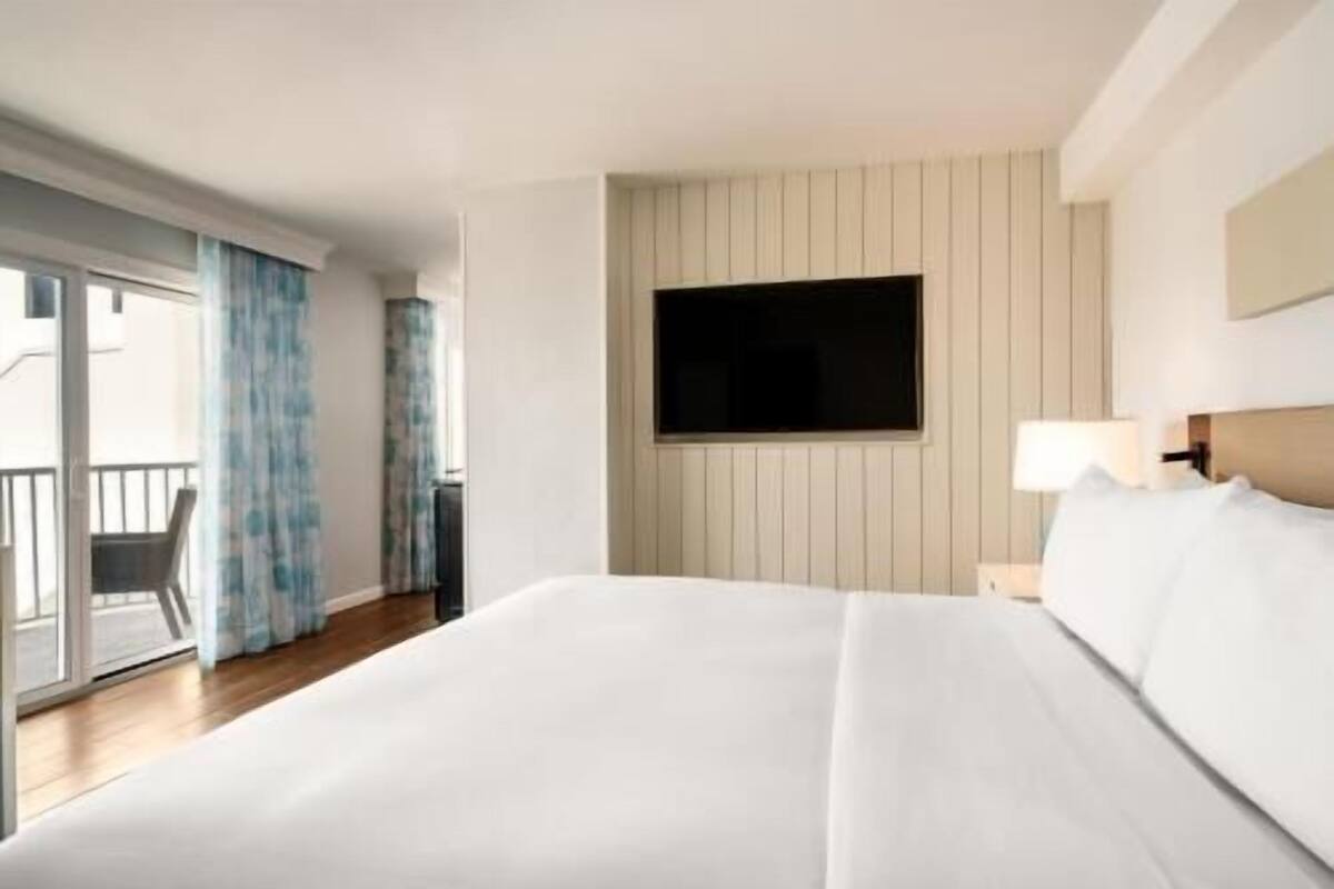A hotel room with a bed, a TV mounted on the wall, a balcony with curtains, and a wooden floor.