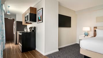 Habitación de hotel con kitchenette, televisión de pantalla plana, cama con edredón blanco y alfombra estampada.