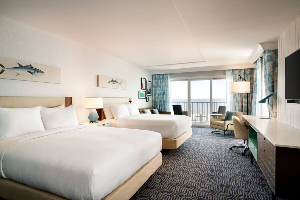 A hotel room with two beds, a desk, a chair, a TV, and a balcony with curtains.