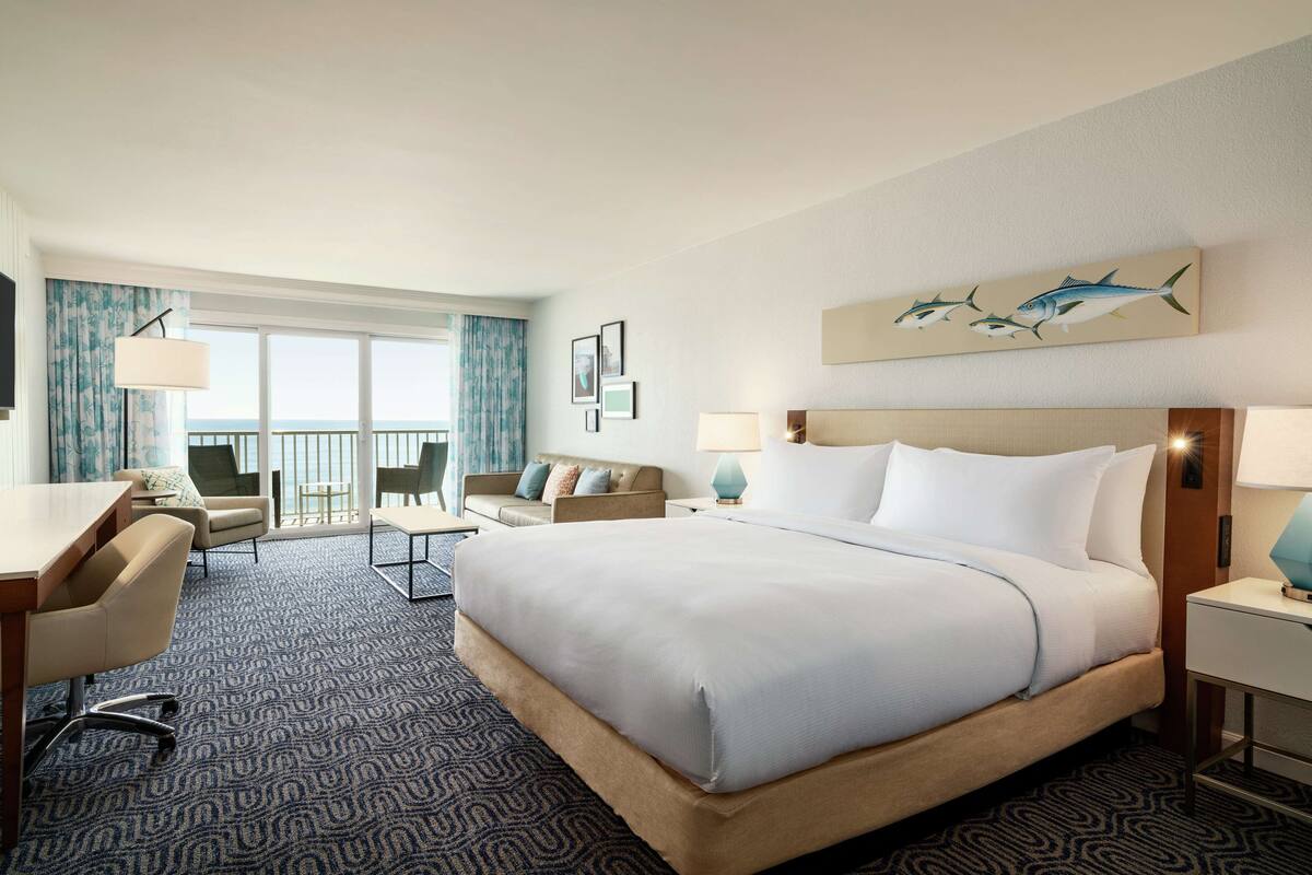 A hotel room with a large bed, a desk, a chair, a sofa, and a balcony with a view.