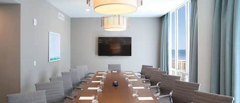 Un salón de conferencias con una mesa larga, sillas, una pantalla de televisión y un ventanal con cortinas.