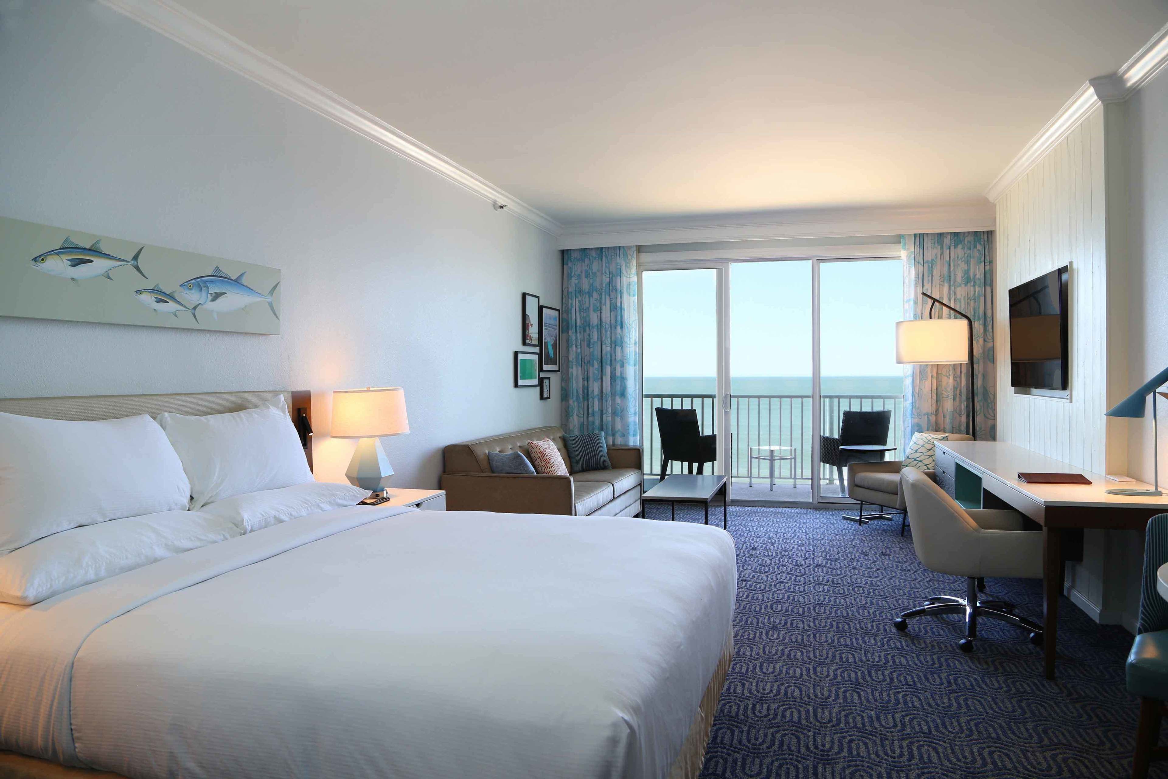 A hotel room with a large bed, a desk, a chair, a sofa, a balcony with a view of the sea, and a painting of fish on the wall.