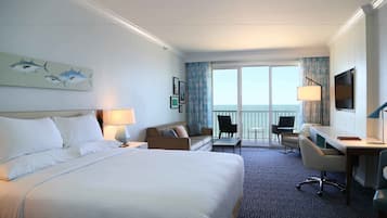 A hotel room with a large bed, a desk, a chair, a sofa, a balcony with a view of the sea, and a painting of fish on the wall.