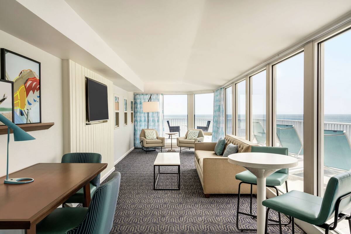 A modern hotel room with a large balcony, a sofa set, a dining table, and a TV.