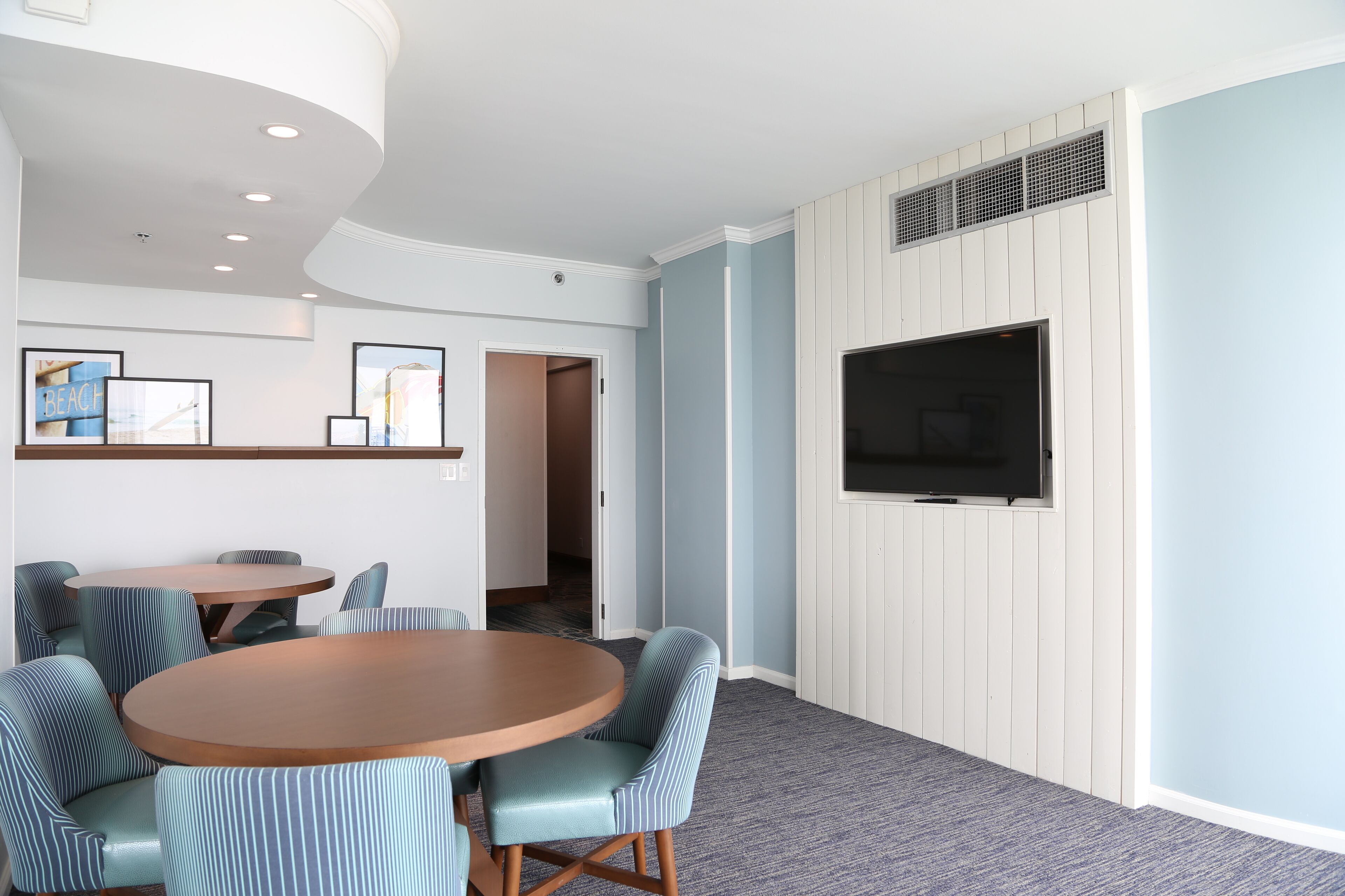 A hotel room with a round wooden table, striped upholstered chairs, a flat-screen TV mounted on a white paneled wall, and framed pictures on the wall.