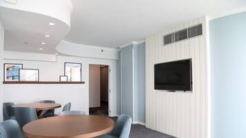 A hotel room with a round wooden table, striped upholstered chairs, a flat-screen TV mounted on a white paneled wall, and framed pictures on the wall.