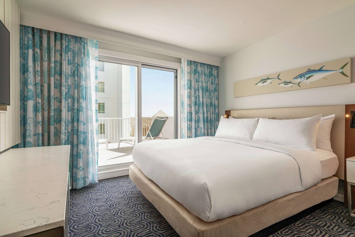 A hotel room with a large bed, a desk, and a balcony with curtains.