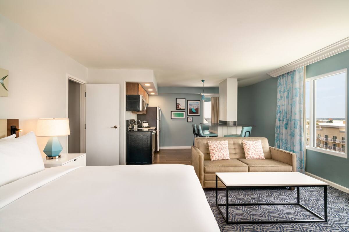 A hotel room with a bed, a desk, a sofa, a kitchenette, and a view of the city.
