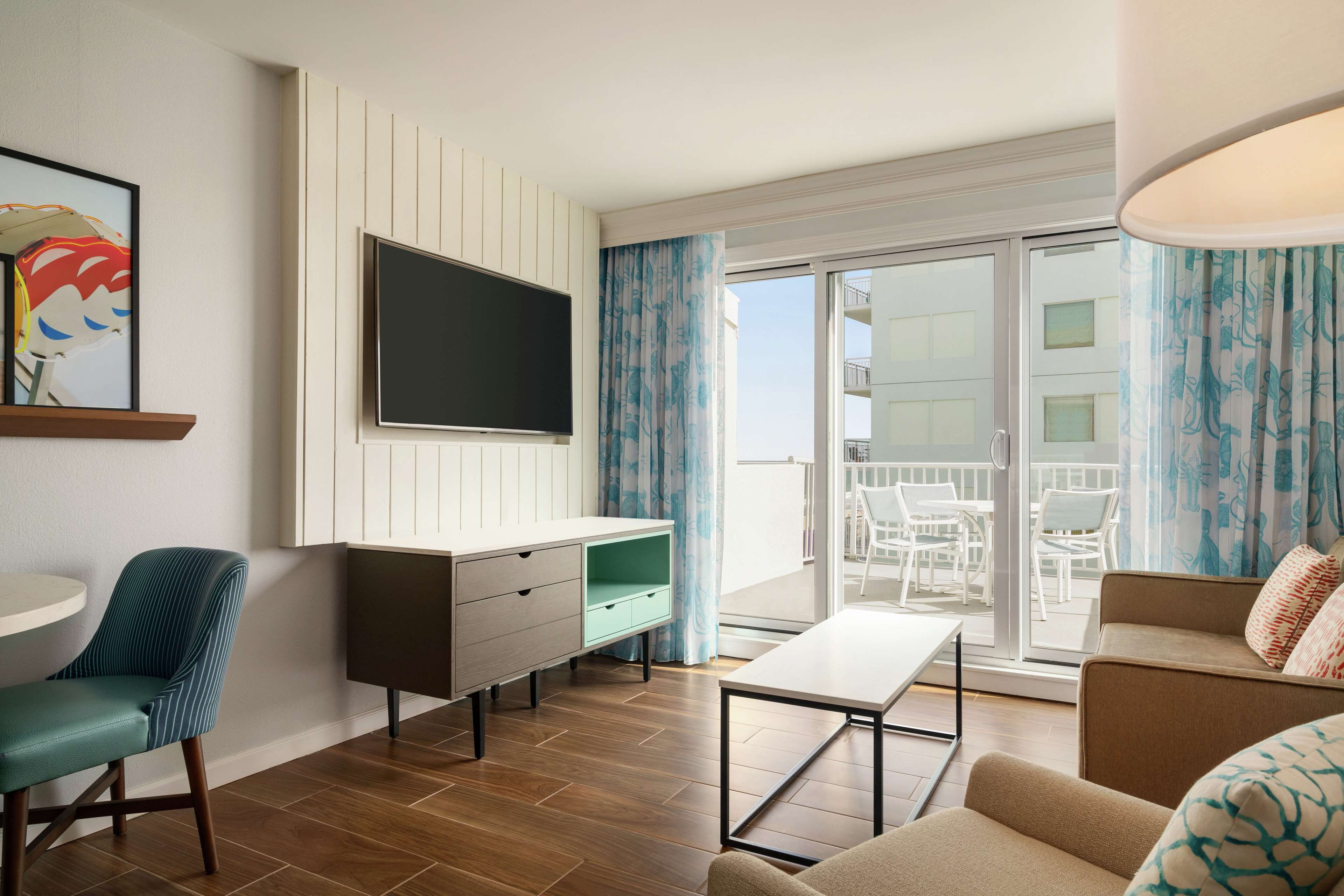 A modern living room with a flat-screen TV, a dining area with a table and chairs, and a balcony with a view of the building.