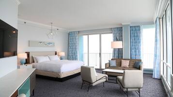 Habitación de hotel con cama, dos sillas, escritorio, televisor y una ventana grande con cortinas.