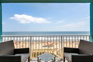 Varanda com móveis de vime e uma mesa, com vista para uma praia com guarda-sóis e pessoas.