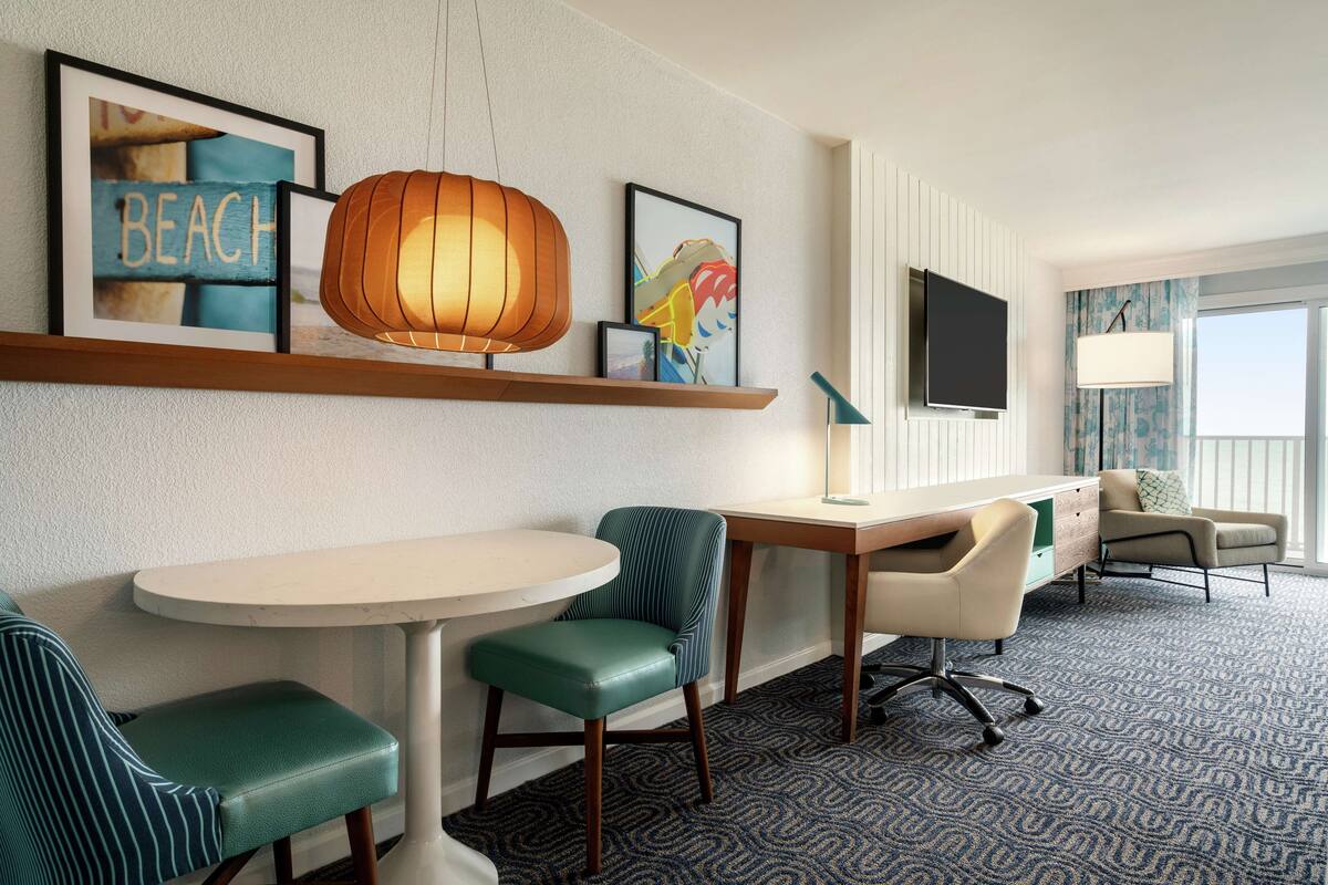 A hotel room with a round dining table, two green upholstered chairs, a wooden desk with a chair, a TV mounted on the wall, and a large window with a view.