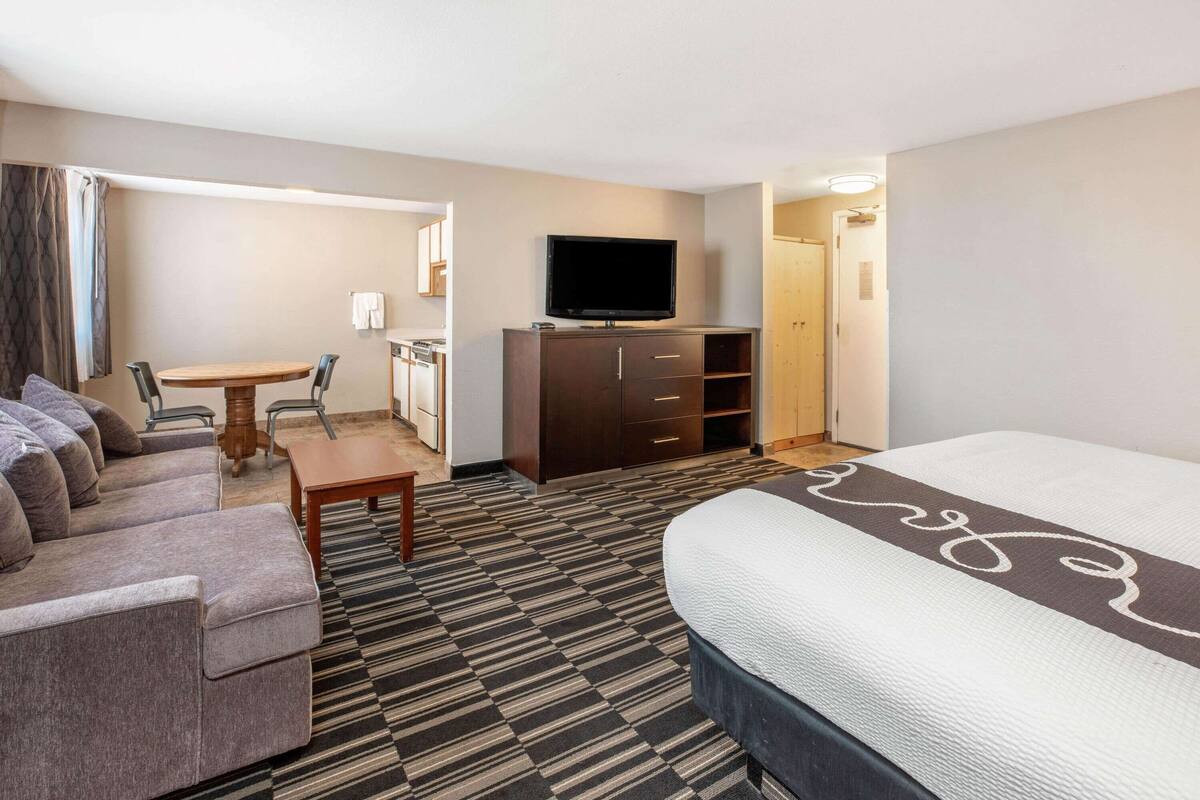 A hotel room with a large bed, a sofa, a small table, a TV on a cabinet, and a kitchenette in the background.