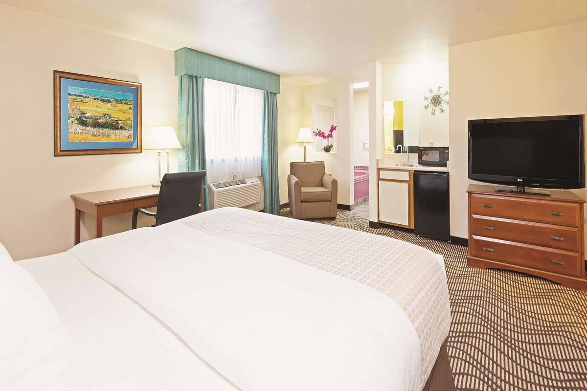 A hotel room with a large bed, a desk with a chair, a television, and a small kitchenette.