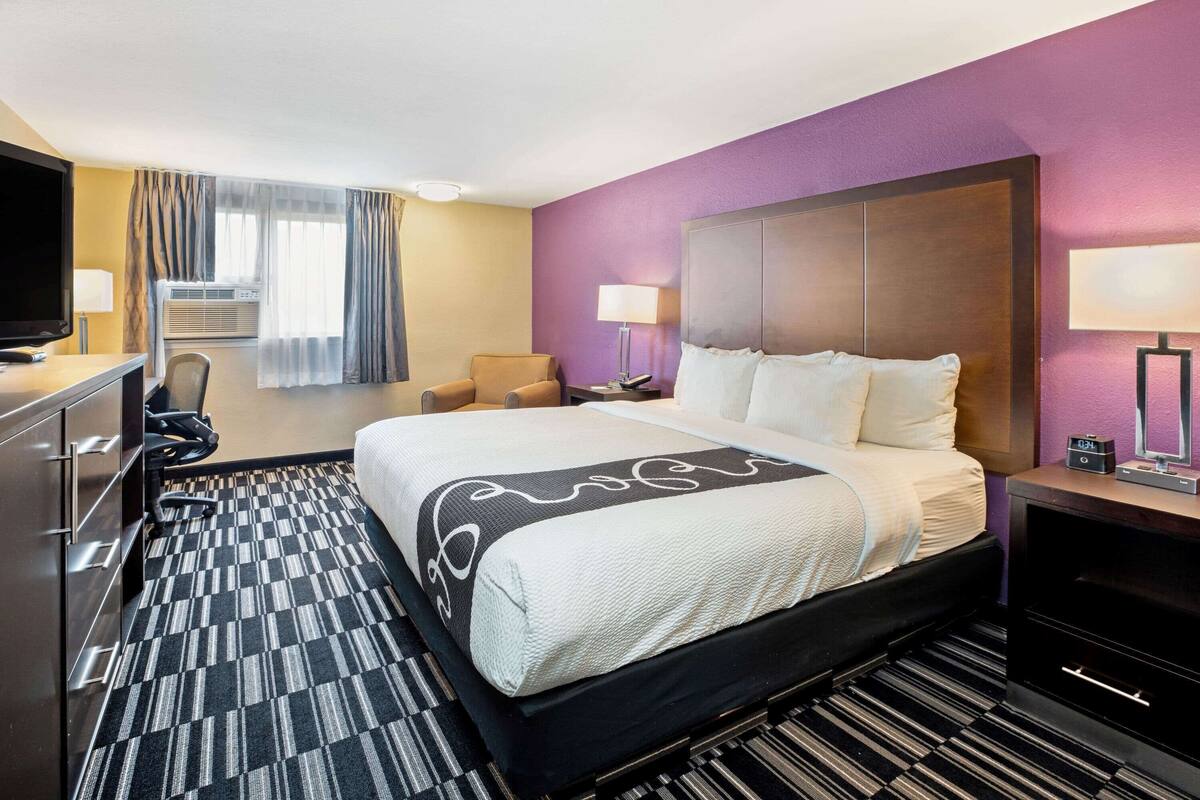A hotel room with a bed, a desk with a TV, a chair, a lamp, and a window with curtains.