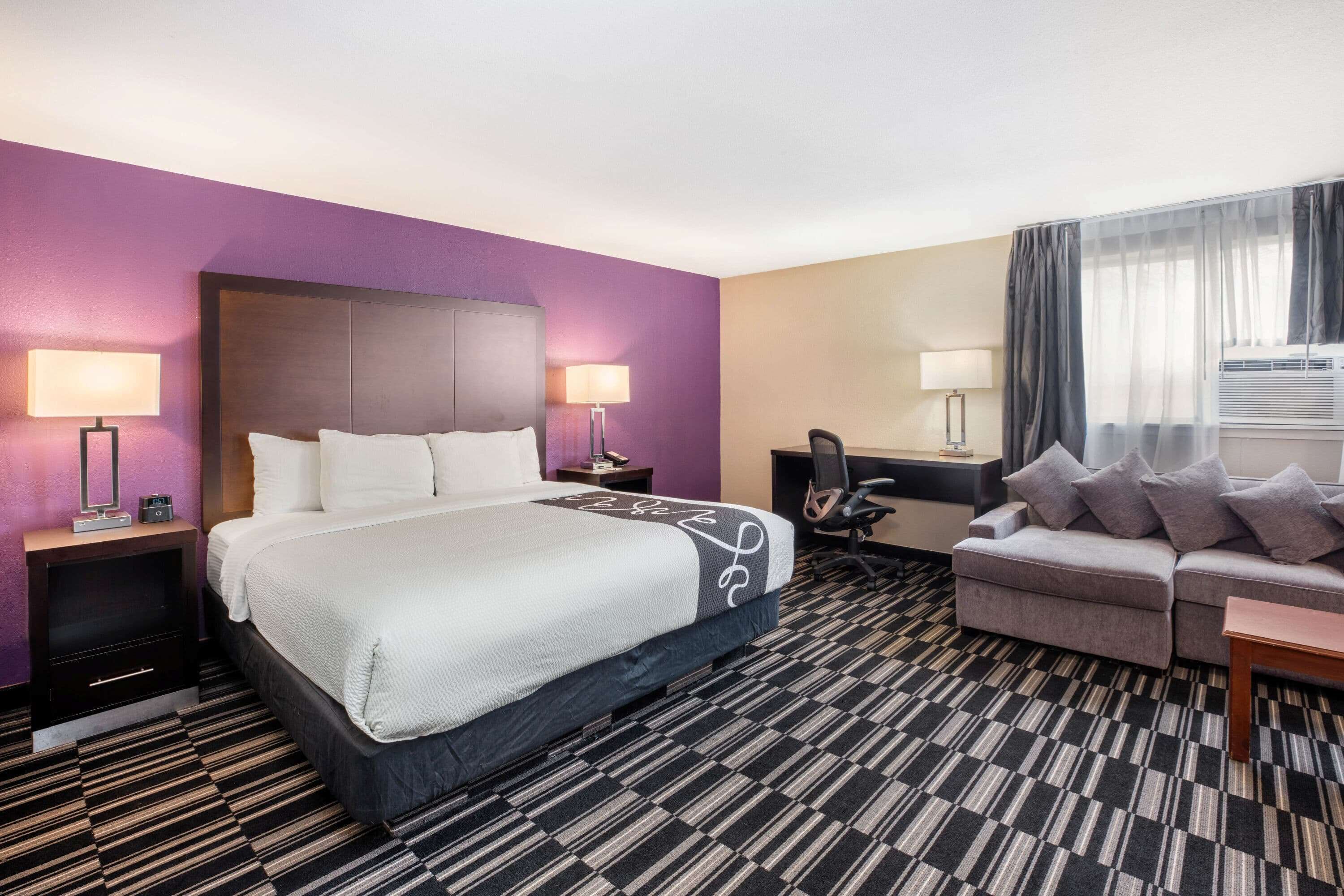 Een hotelkamer met een groot bed, een bank, een bureau en een stoel. Er staan lampen aan beide zijden van het bed en er is een raam met gordijnen.