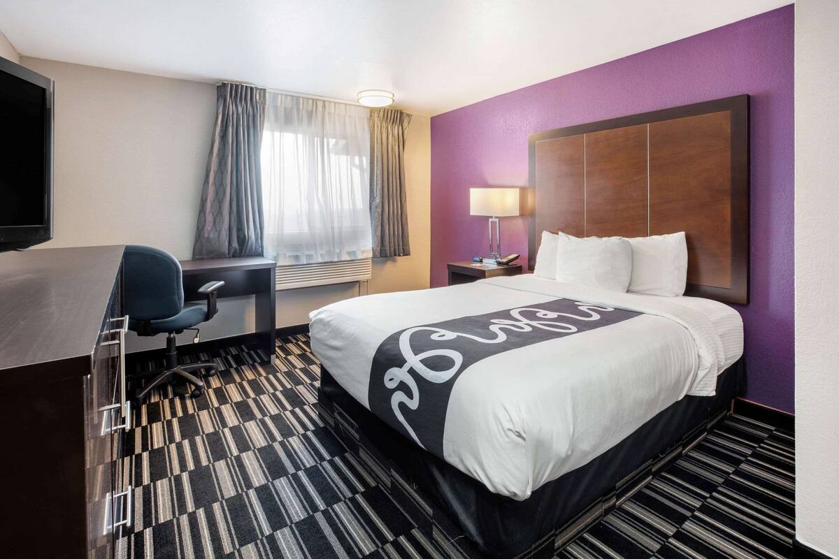 A hotel room with a bed, a desk, a chair, a TV, a window with curtains, and a nightstand with a lamp.