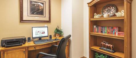 A hotel room with a wooden desk, a computer, a printer, a bookshelf, and a framed picture on the wall.