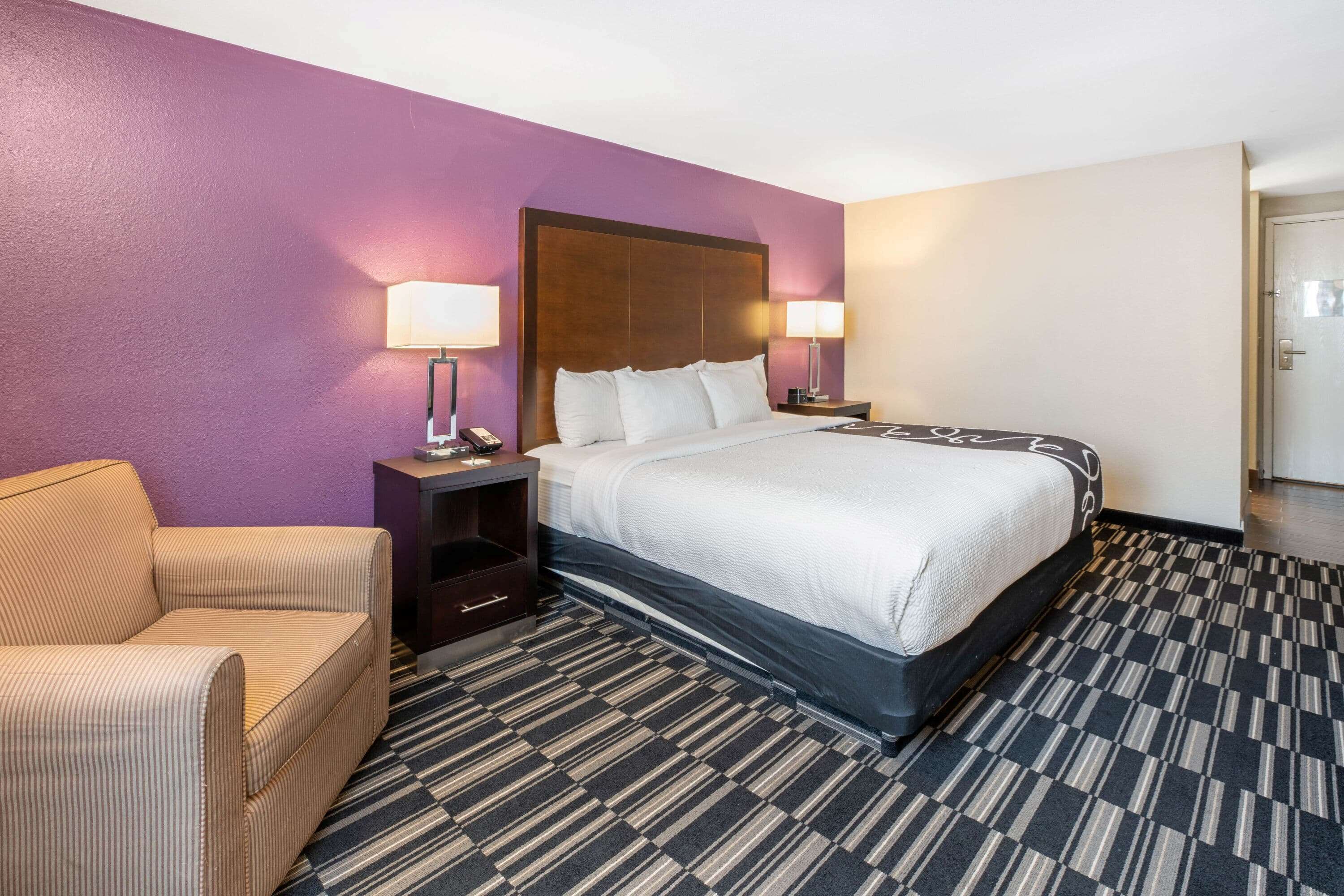 A hotel room with a large bed, two bedside tables with lamps, a chair, and a patterned carpet.