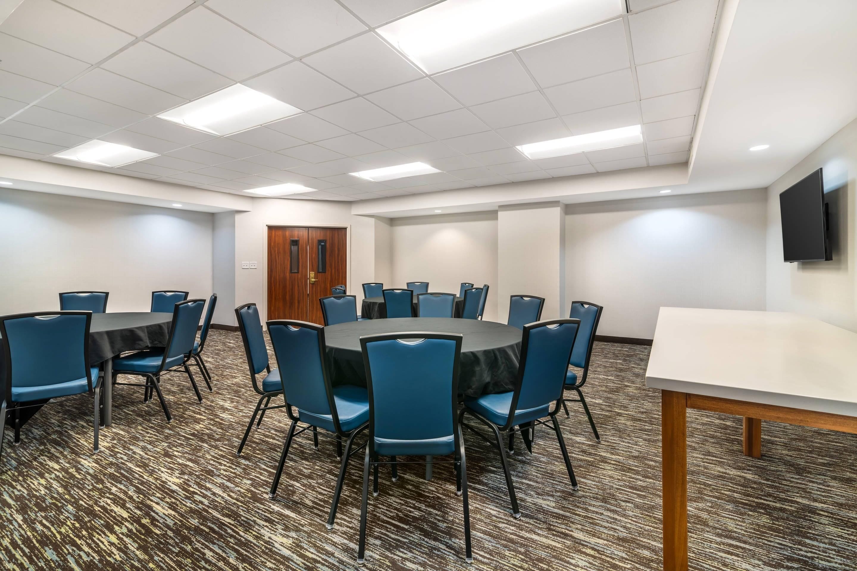 A conference room with a round table and blue chairs, carpeted floor, white walls, and a wooden door.