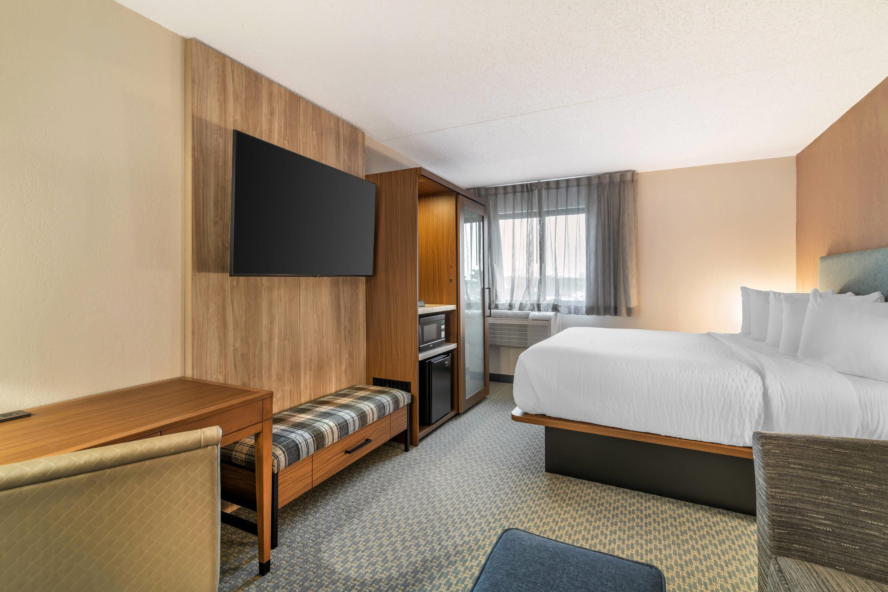 Habitación de hotel con cama, escritorio, televisión, zona de estar y ventana con cortinas.