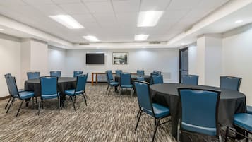 Un salón de conferencias con mesas redondas y sillas azules, suelo alfombrado, paredes blancas, un televisor de pantalla plana y una puerta.