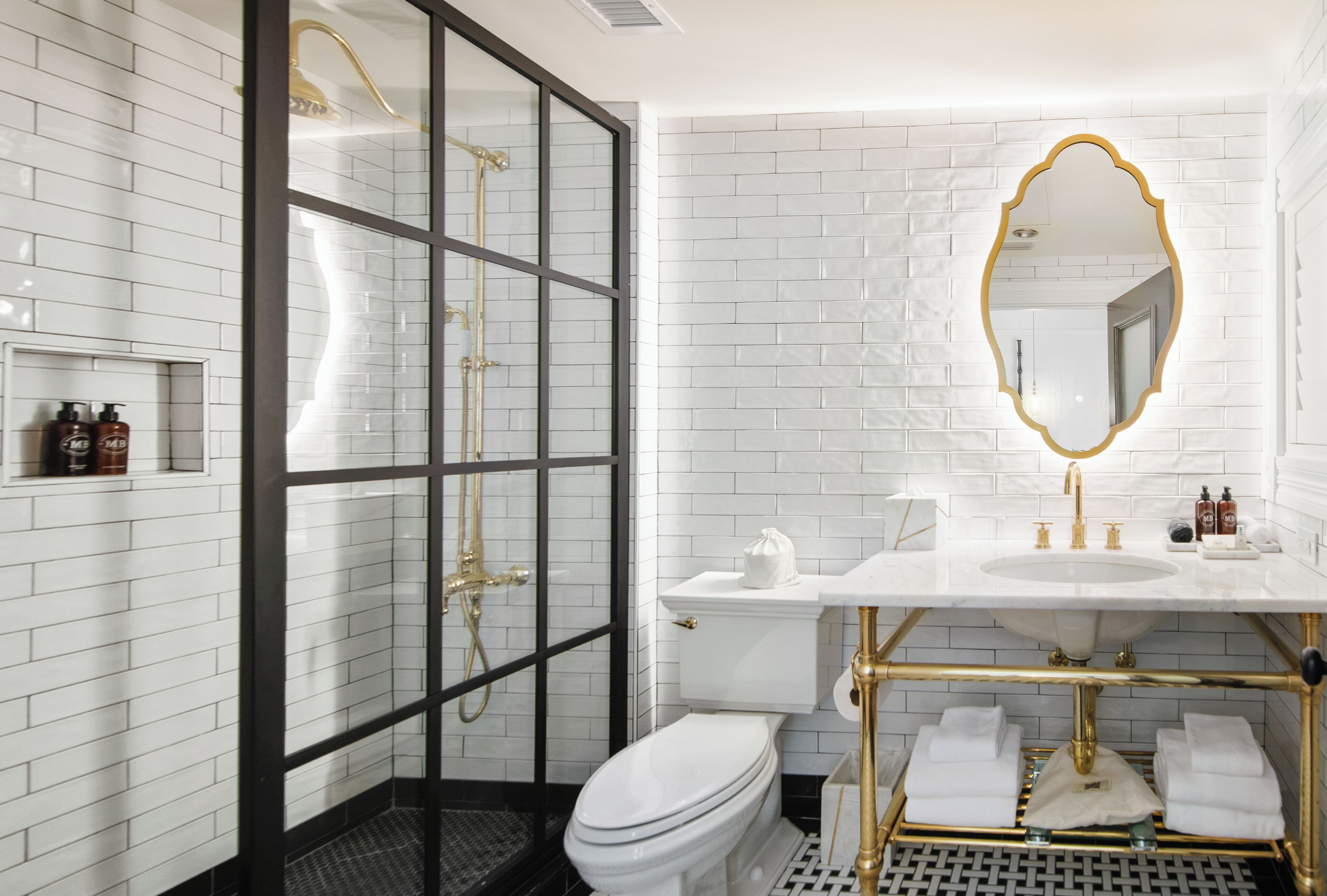 A bathroom with a white-tiled wall, a glass-enclosed shower, a gold-framed mirror, and a white sink with gold fixtures.