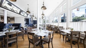 A spacious dining area with multiple tables and chairs, large windows offering views of the city, and a brick wall in the background. - Fairmount Hotel (San Antonio)