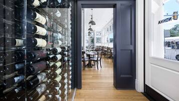 A wine cellar with bottles on shelves, a wooden floor, and a view of a dining area with tables and chairs.