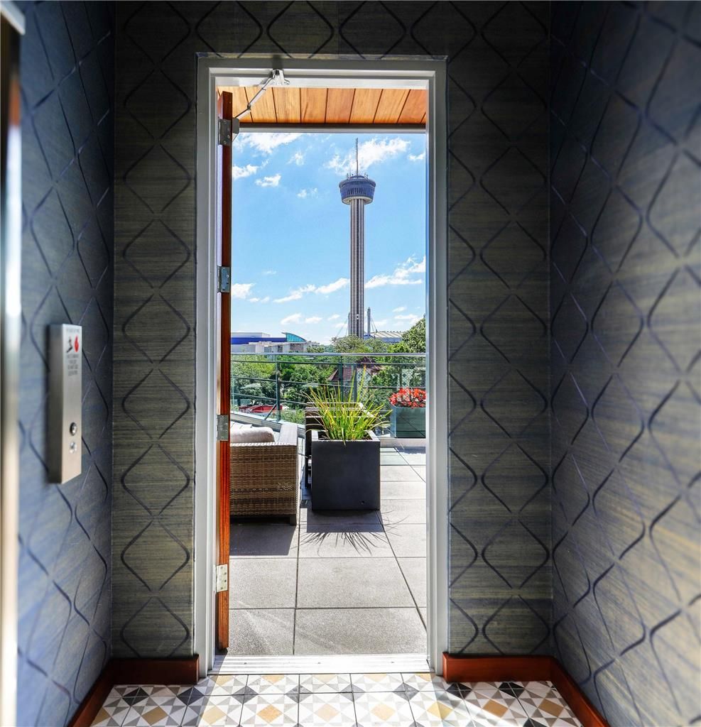 Vista a través de una puerta que da a un espacio exterior con suelo de baldosas, una planta en maceta y una silla de mimbre.