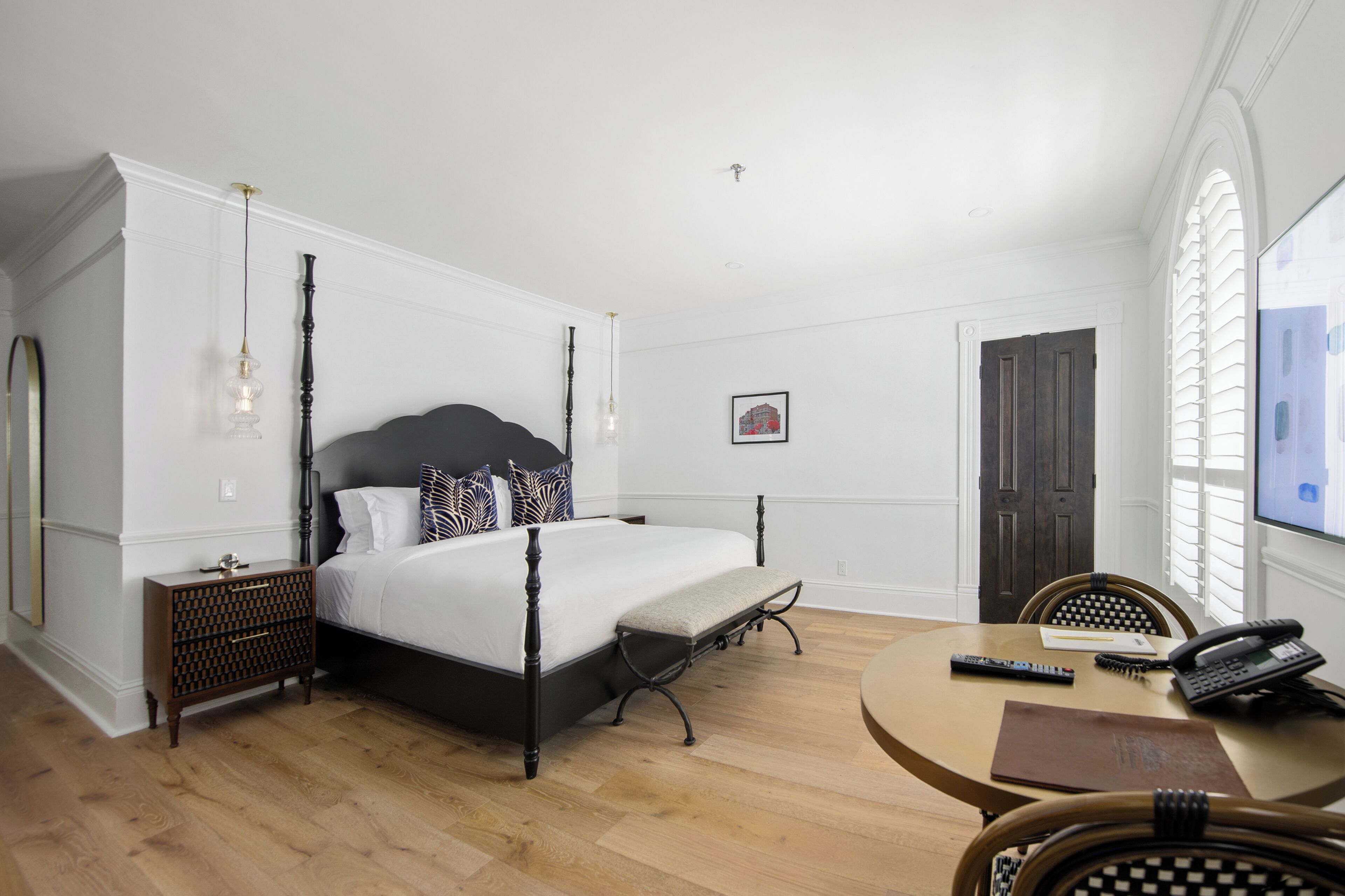 A bedroom with a four-poster bed, a bedside table, a chair, a round table with a phone and remote control, and a window with blinds.