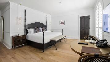 A bedroom with a four-poster bed, a bedside table, a chair, a round table with a phone and remote control, and a window with blinds.
