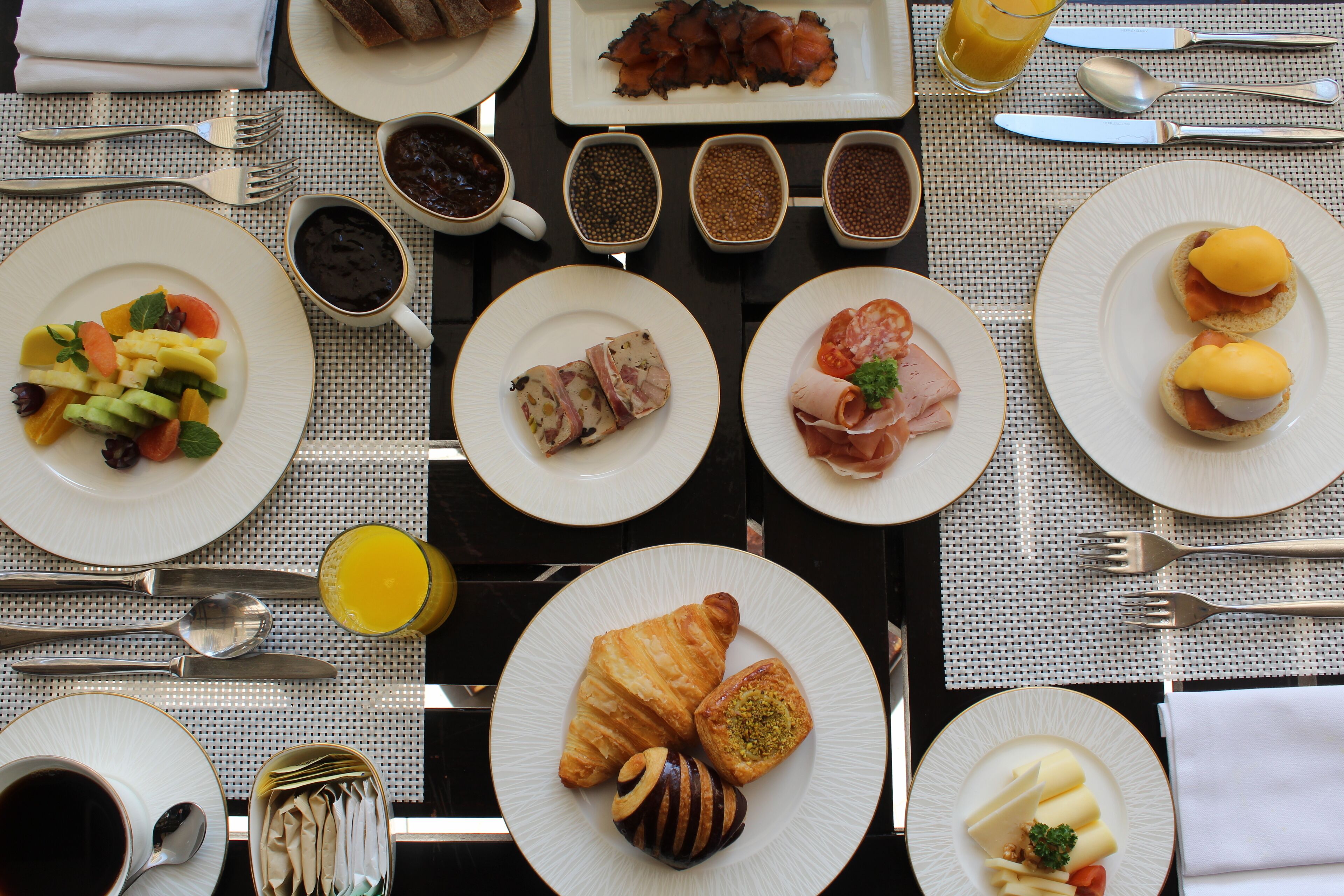 Buffet de café da manhã com diversos pratos, incluindo frutas, carnes e doces, disposto sobre uma mesa com toalha xadrez.
