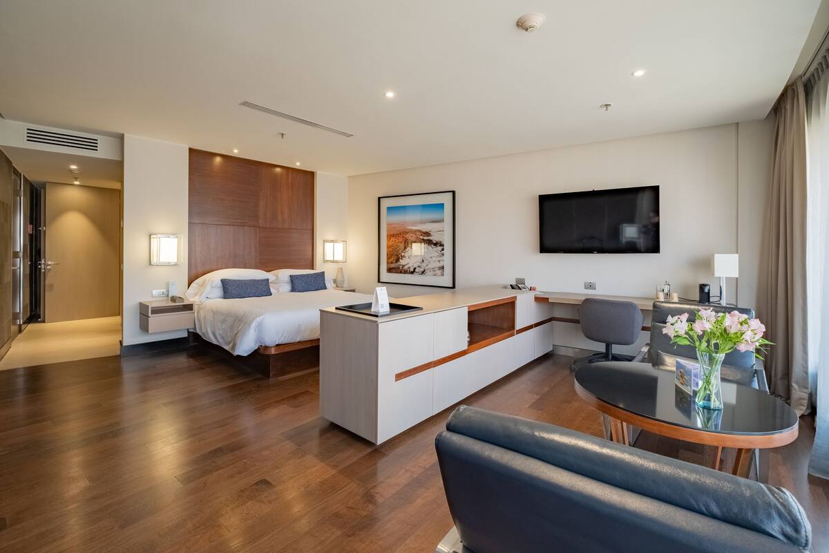 A modern hotel room with a bed, a desk, a TV, and a seating area.