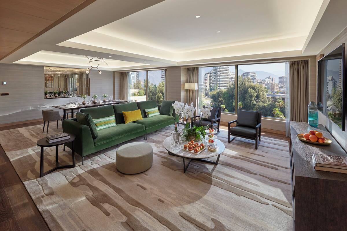 A modern living room with a green sofa, a round coffee table with fruit, and a city view through large windows.