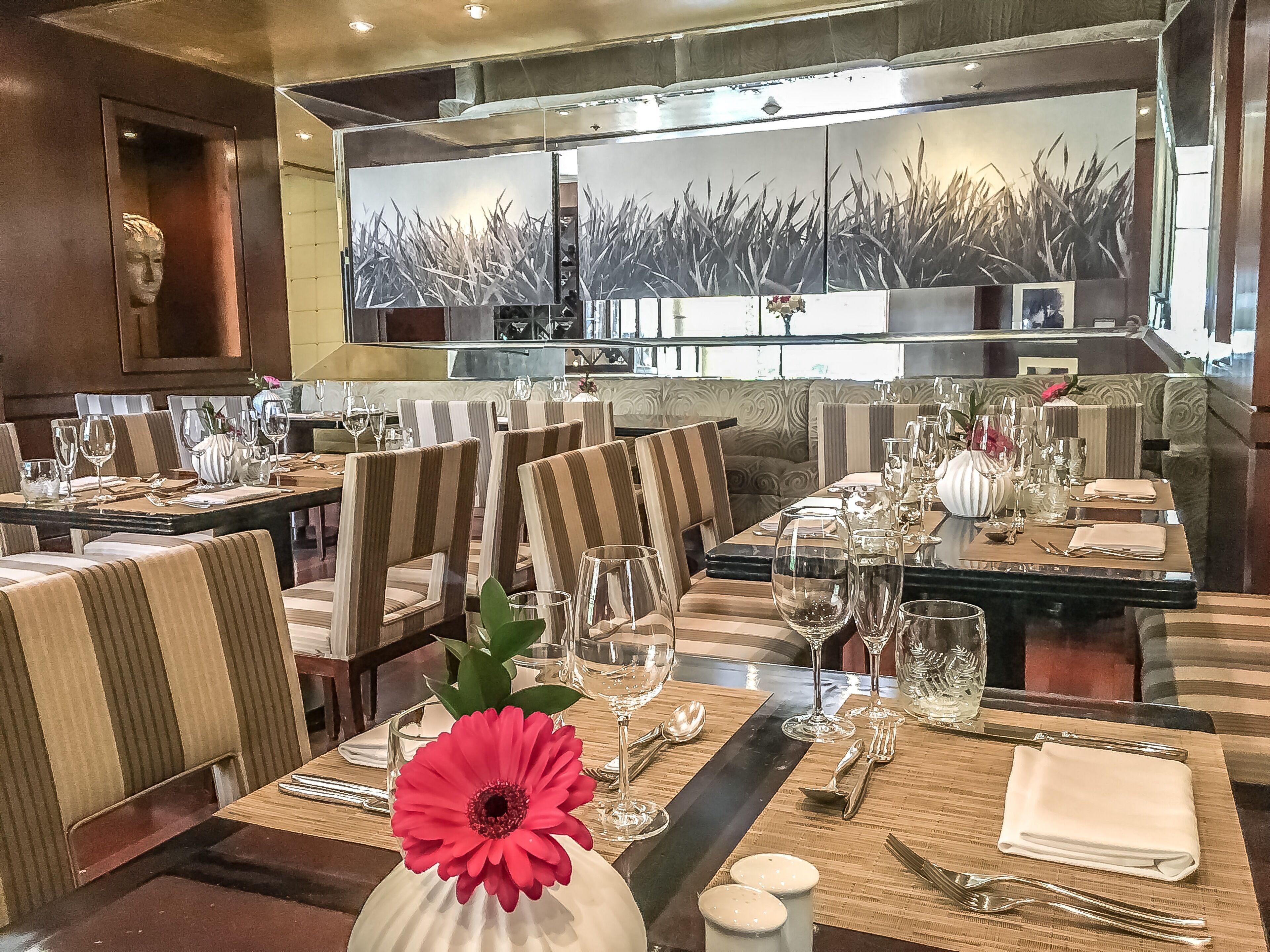 A restaurant with striped upholstered chairs, neatly set dining tables with wine glasses, and a large mirror reflecting the room.