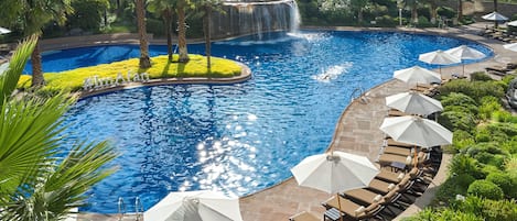 A swimming pool with a waterfall feature, surrounded by palm trees and lounge chairs with umbrellas.