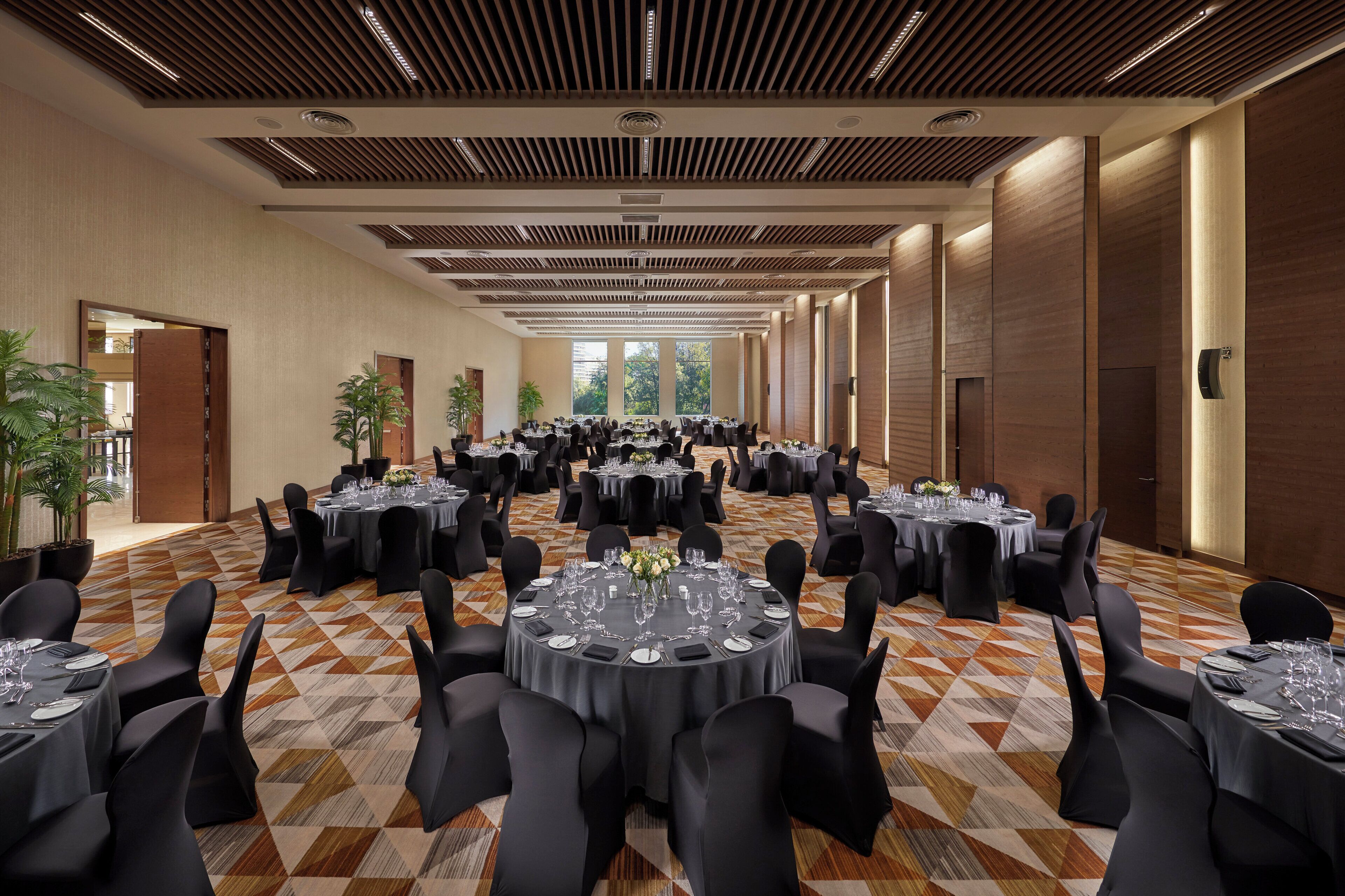Uma grande sala de banquetes com várias mesas redondas dispostas para refeições, apresentando um tapete com padrão geométrico e teto de painéis de madeira.