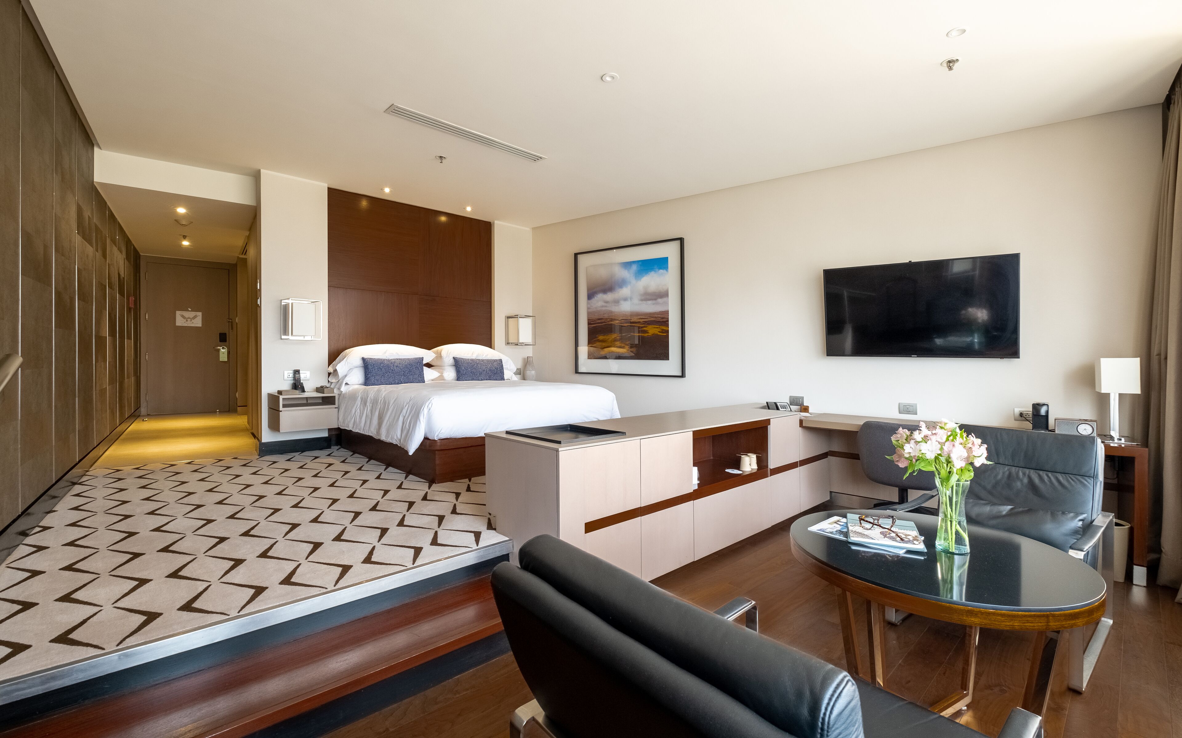Quarto de hotel com cama, escrivaninha, cadeira, televisão e um vaso com flores.