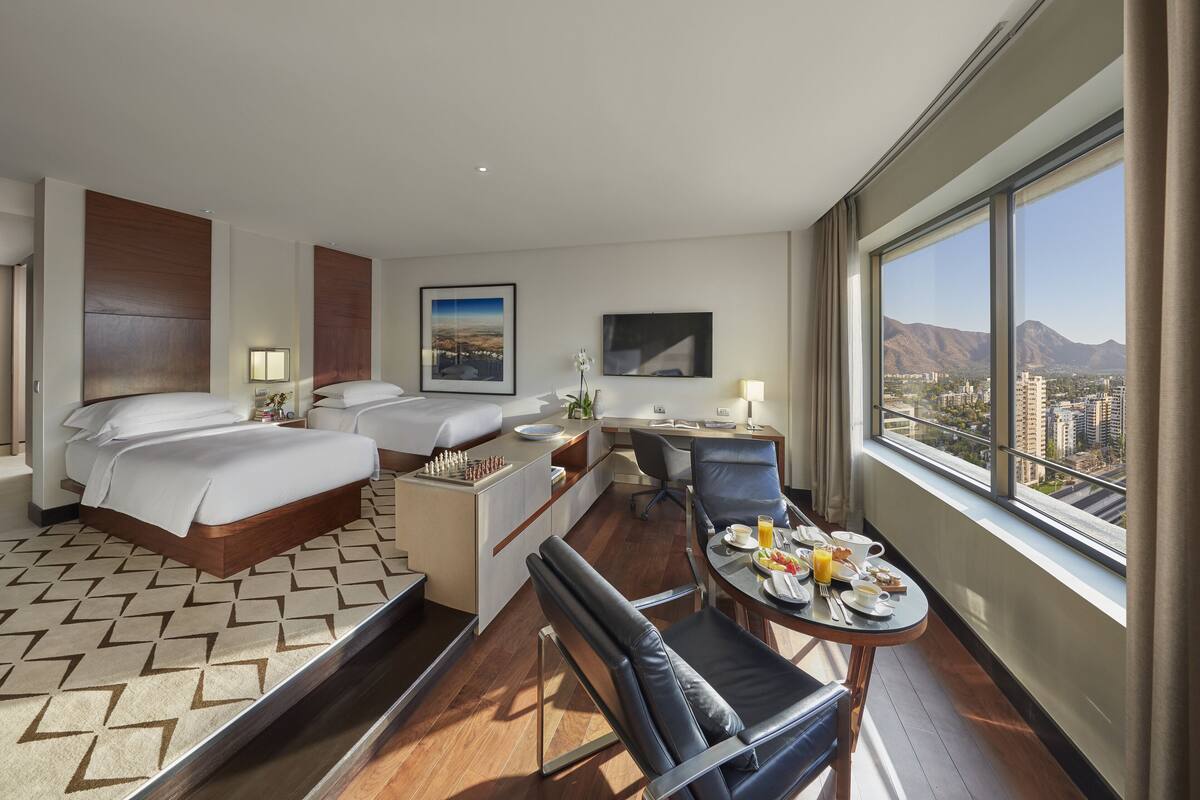 A hotel room with two beds, a desk, a dining area, and a large window with a city and mountain view.