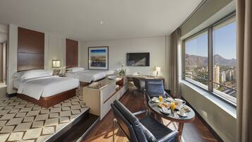A hotel room with two beds, a desk, a dining area, and a large window with a city and mountain view.