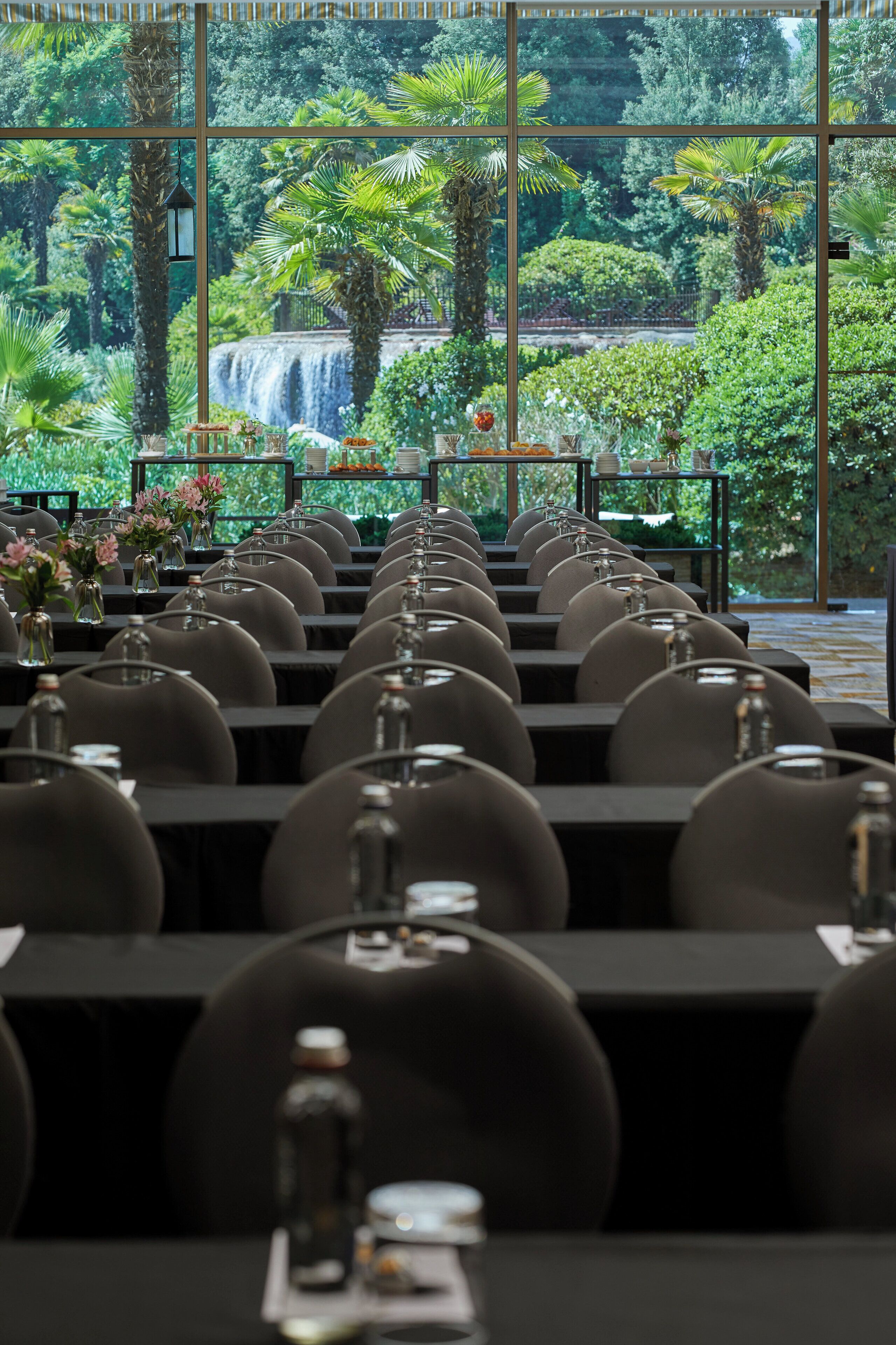 Un restaurante con filas de mesas y sillas, vistas a una cascada y un jardín tropical.