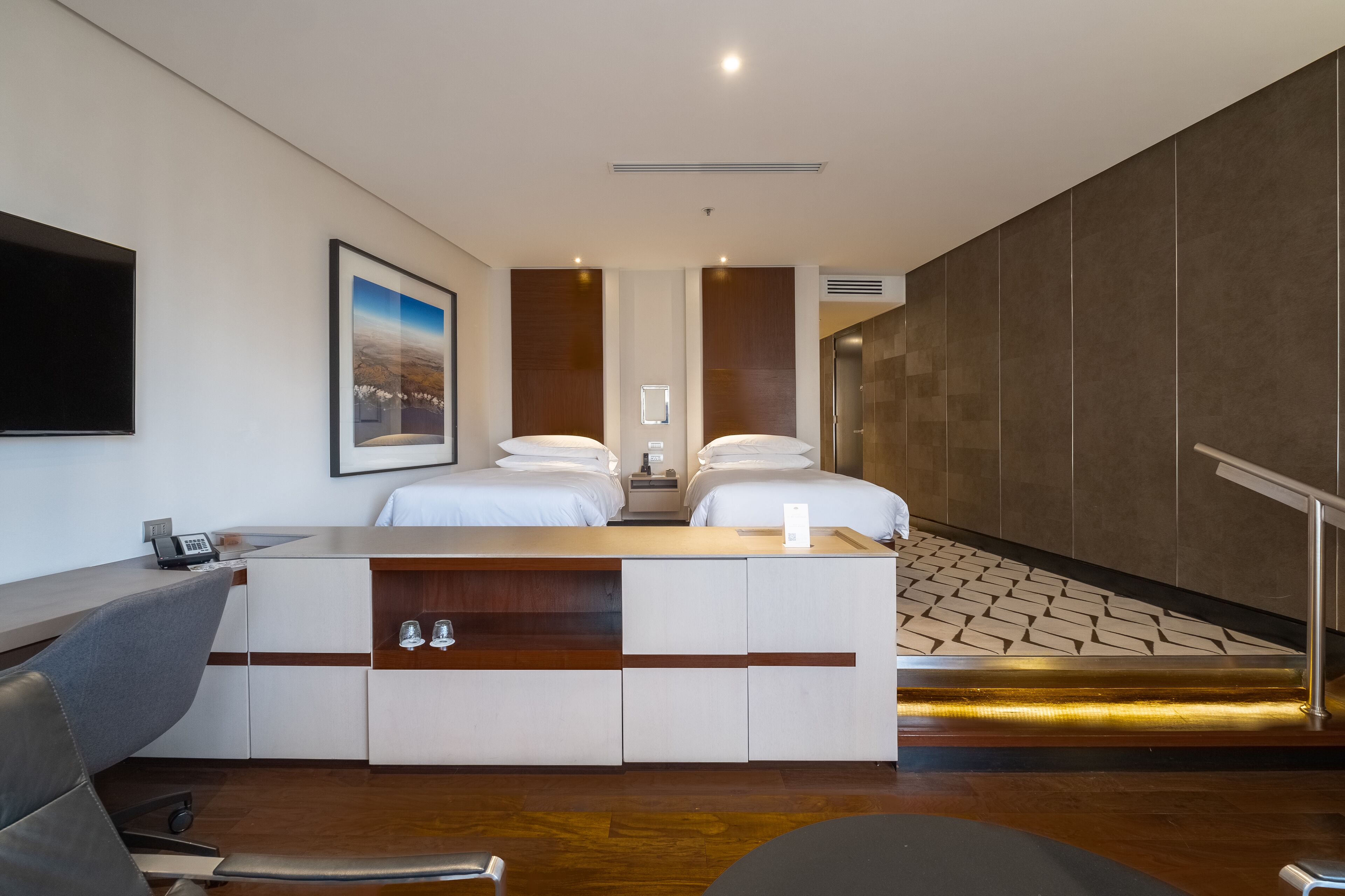 A hotel room with a desk, two beds, a TV, a framed picture, and a staircase.
