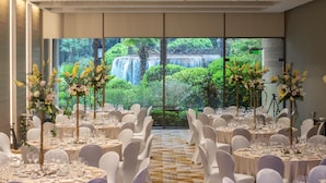 Un salón de banquetes con mesas redondas, sillas blancas y centros de mesa florales. Se ve una cascada a través de amplias ventanas.