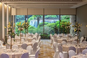 Un salón de banquetes con mesas redondas, sillas blancas y centros de mesa florales. Se ve una cascada a través de amplias ventanas.