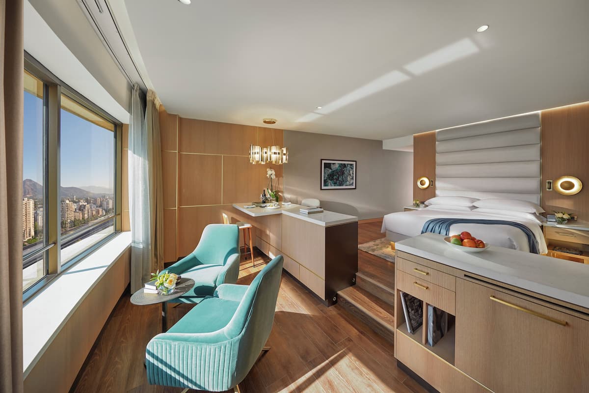 Una habitación de hotel moderna con un ventanal grande, un escritorio, dos sillas, una cama y una pequeña kitchenette.