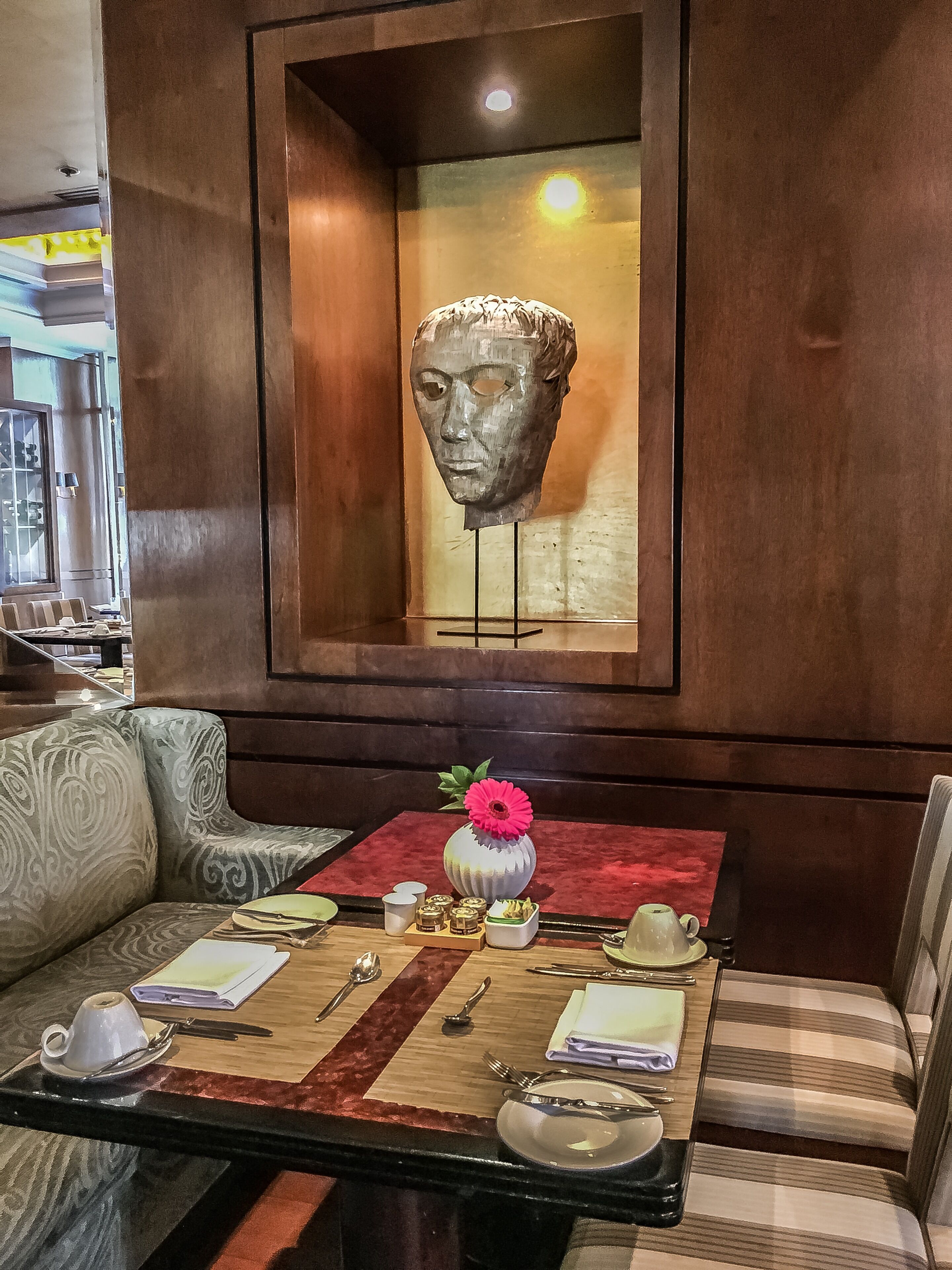 Un coin repas avec une table dressée pour deux personnes, un vase avec une fleur et une sculpture en buste accrochée au mur.